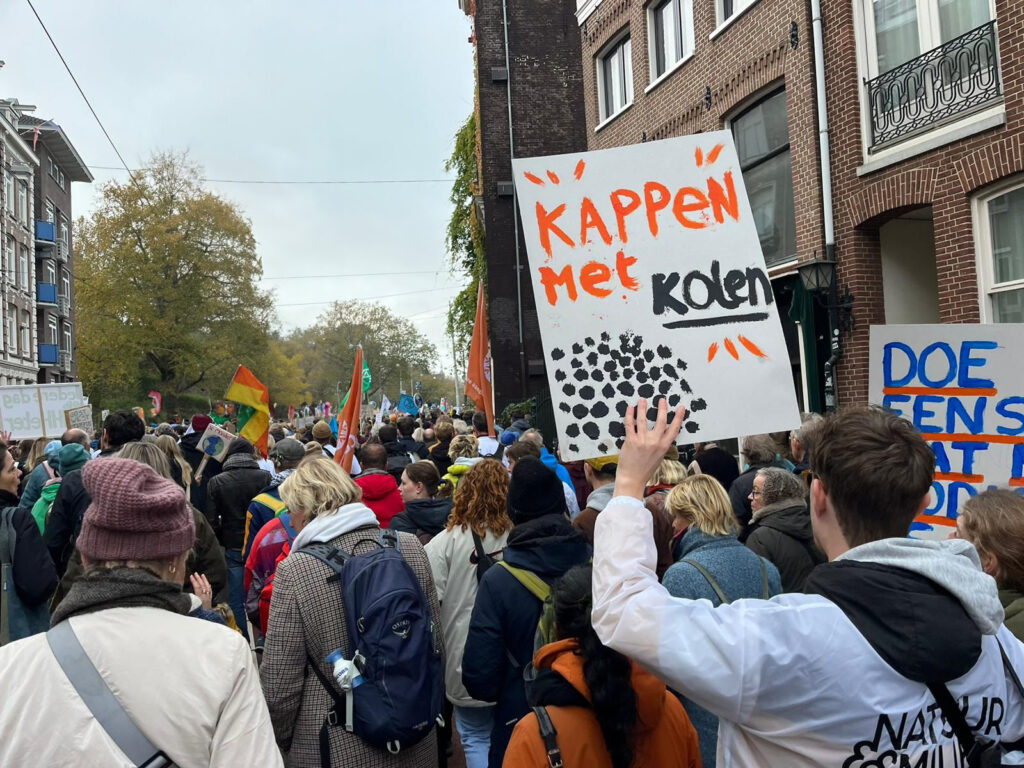 Menigte mensen die meelopen met een klimaatmars. Op de voorgrond een man met een bord waarop staat: Kappen met kolen.