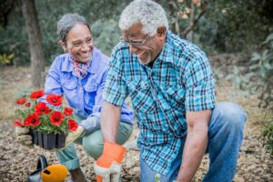 Twee oudere mensen lachen terwijl ze samen in een tuin bloemen planten; ze dragen tuinhandschoenen en knielen tussen bladeren en planten