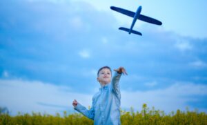 Persoon die een blauw speelgoedvliegtuig de lucht in gooit in een veld met gele bloemen onder een blauwe lucht met wolken (luchtvervuiling)