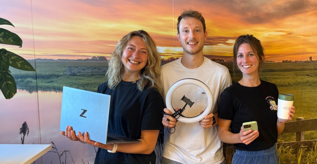 Contentmarketeers Naomi, Mark en Marije met laptop, ringlight en telefoon in de hand tegen een fotowand van een ondergaande zon over een weids landschap