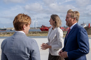 Zijne Majesteit de Koning en Marjolein Demmers, directeur van Natuur & Milieu, in gesprek tijdens het werkbezoek aan De Rijke Noordzee en Ecowende in IJmuiden, met windmolens en schepen in de haven op de achtergrond.