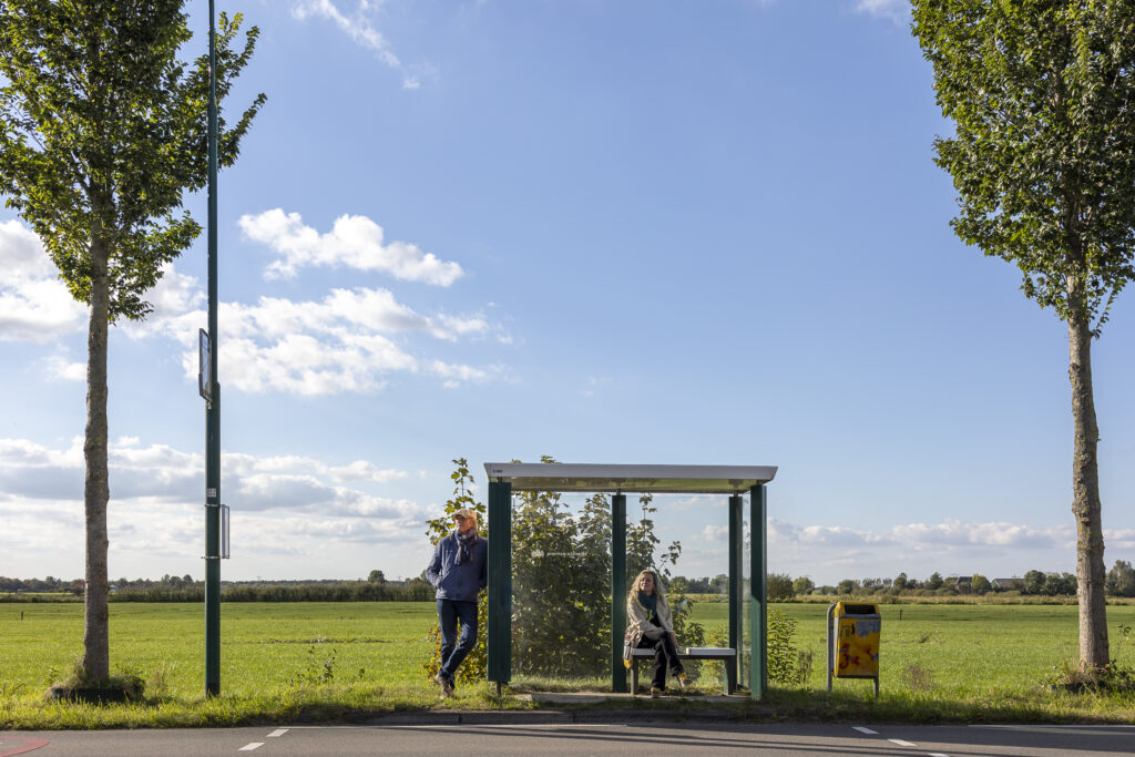 Twee mensen wachten op de bus bij een halte in een landelijk gebied met weilanden op de achtergrond