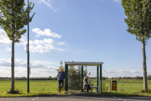 Twee mensen wachten op de bus bij een halte in een landelijk gebied met weilanden op de achtergrond