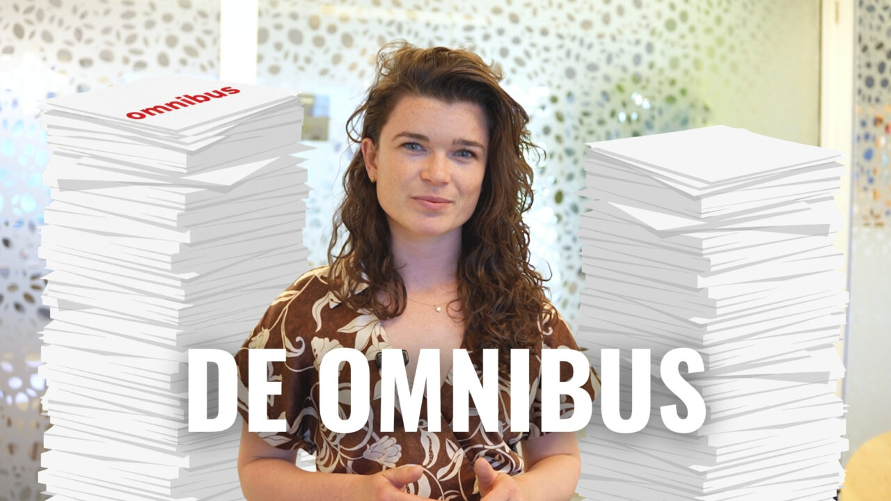 Collega Emmie kijkt de camera in. Achter haar stapels papier en de tekst: 'De Omnibus'