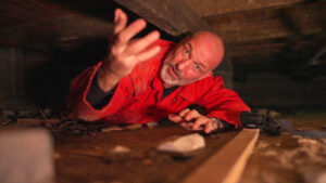 Eddy Zoëy ligt in een rode overall in de kruipruimte onder de vloer