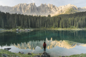 Vrouw staat aan Lago di Carezza in Italië met bergen op de achtergrond die weerspiegelen in het heldere water