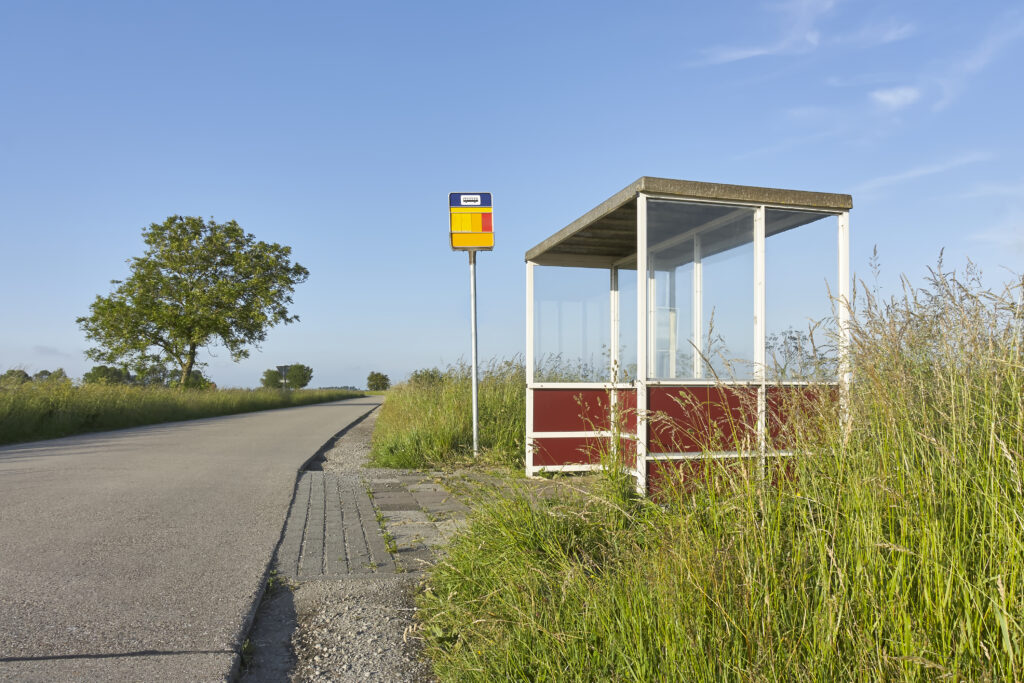 Bushokje met glas langs een weg in een landelijk gebied onder een blauwe lucht.