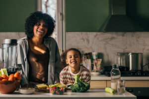 Vrouw staat met een kind in de keuken. Er liggen groenten op het aanrecht.
