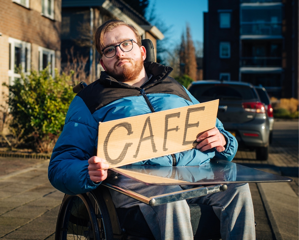 man in rolstoel staat naast de weg te liften met een bord met daarop het woord café